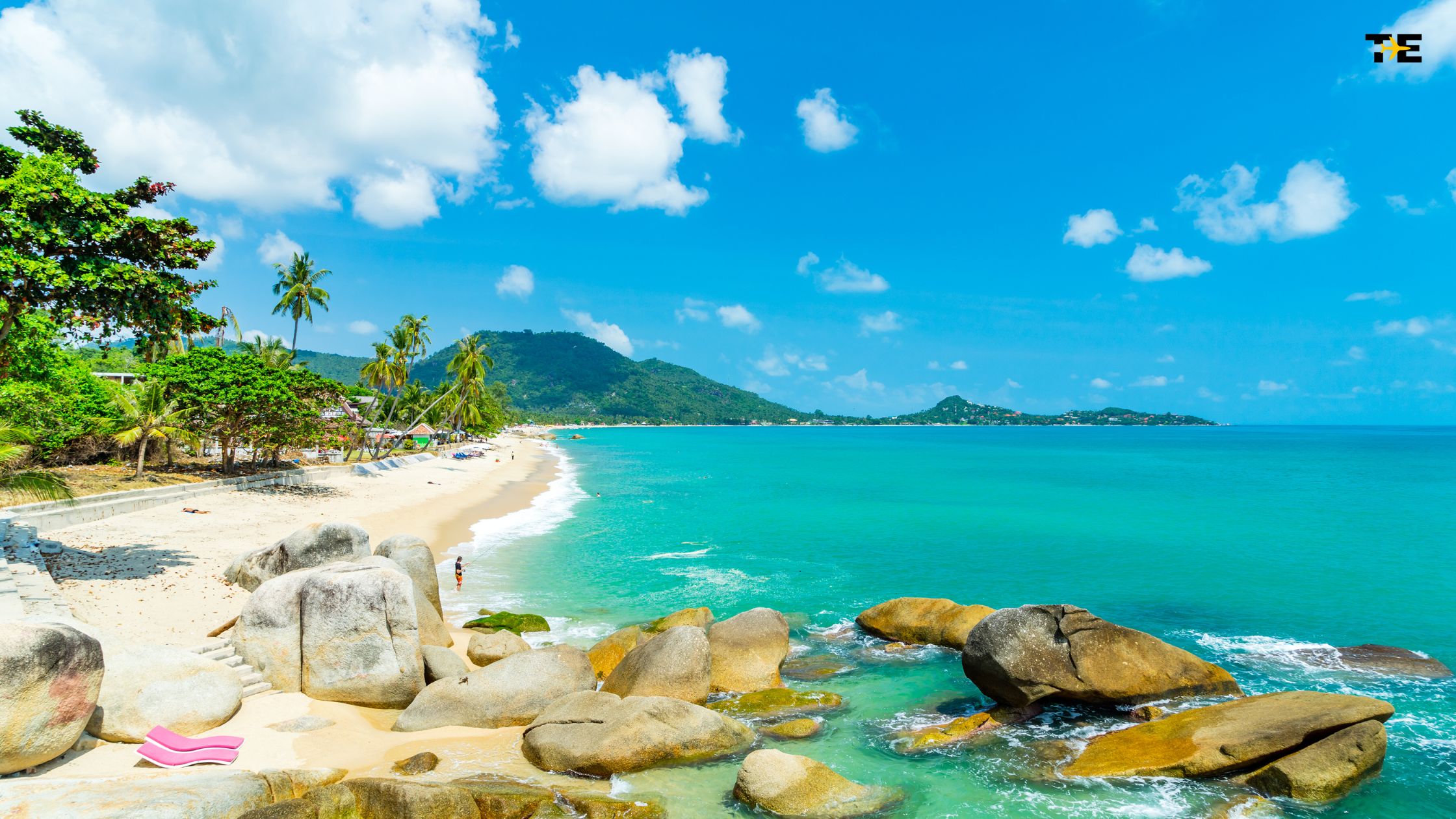 Tropical beach scene with rocks, palm trees, and vibrant loungers under a bright blue sky.