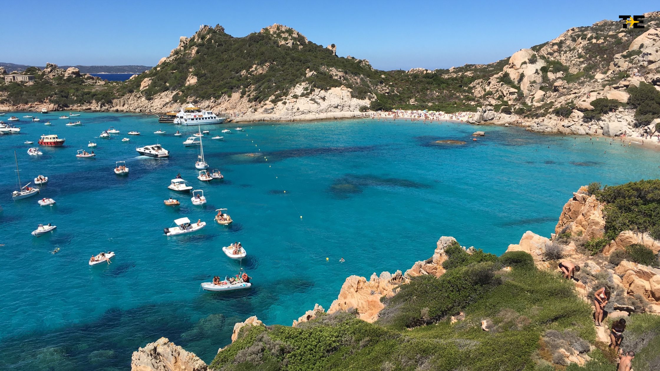 Vibrant coastal bay scene with turquoise waters, boats, beachgoers, and lush green hills.