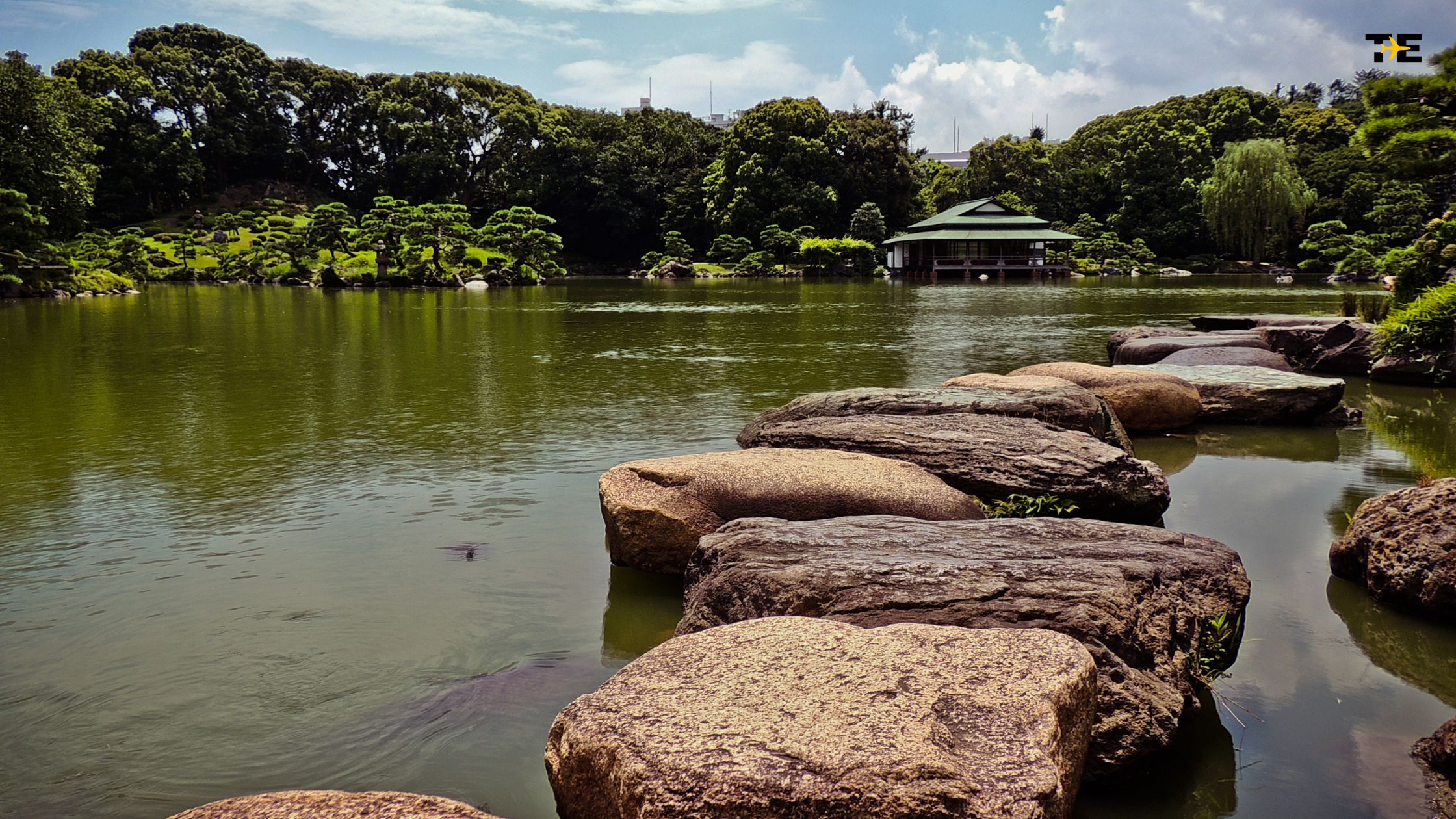 Serene pond pathway with smooth stones surrounded by lush greenery and a traditional structure.